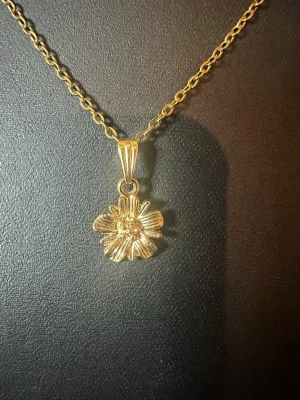 Guldhalsband med blomma - Snyggt halsband i guldfärg med en detaljrik blomformad berlock. Kedjan är tunn och elegant, och blomman har fina räfflade blad som ger ett unikt intryck. Perfekt accessoar för dig som gillar stilrena och feminina smycken.