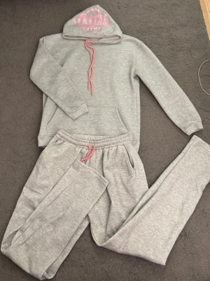 Synaworld set - Mysigt grått loungewear-set med hoodie och byxor. Hoodien har rosa dragsko och tryck på luvan, samt magficka. Byxorna har resår i midjan och rosa dragsko. Perfekt för chill dagar hemma.