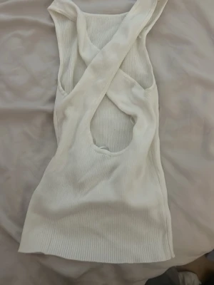 Vit ribbad linnetopp med öppen rygg - Säljer en vit ribbad linnetopp i stretchigt material. Toppen är ärmlös och har en snygg öppen rygg med korsade band som ger en unik look. Perfekt till sommarens outfits och lätt att matcha med olika stilar.