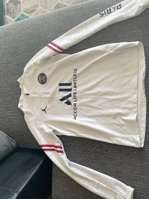 Vit PSG träningströja Jordan - Snygg vit långärmad träningströja från PSG och Jordan med halv dragkedja. Tröjan har blå PSG-logga på bröstet, Jordan-logga och texten 'Accor Live Limitless' framtill. Röda och svarta ränder på ärmarna ger extra stil. Perfekt för fotbollsträning eller streetwear.