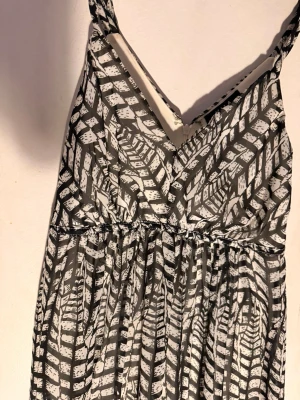 Svartvit långklänning med mönster - Snygg svartvit långklänning med grafiskt mönster och smala axelband. Klänningen har omlott upptill och ett lätt, luftigt tyg som ger ett flowy fall. Perfekt för dig som gillar en unik och trendig stil. Klänningen har underkjol. 