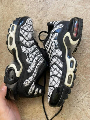 Nike Air Max Plus Tn svart/vit - Nike Air Max Plus Tn sneakers i svart och vitt med rutigt mönster på ovandelen. Skorna har synliga Air-enheter i sulan, svarta detaljer och vita inslag längs sidorna. Ikonisk Tn-logga på sulan och klassisk sportig siluett. Perfekta för dig som gillar streetwear.