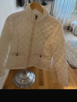 Vit quiltad vindjacka med hög krage - Stilren vit vindjacka med quiltat mönster och hög krage. Jackan har dragkedja framtill, två fickor med knappdetaljer och lång ärm. Perfekt för dig som vill ha en lätt och snygg jacka till vardagslooken.