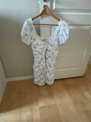 Vit blommig kortklänning med puffärm - Supersöt vit kortklänning med blått blommönster och puffärmar. Klänningen har rynkade detaljer framtill och en feminin, figurnära passform. Perfekt för sommarens alla tillfällen!