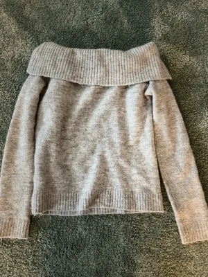 Ljusgrå off shoulder stickad tröja - Mysig ljusgrå stickad tröja med bred ribbad off shoulder-krage och långa ärmar. Tröjan har ribbade muddar vid ärmslut och nederkant, perfekt för lager på lager och en avslappnad men trendig look.