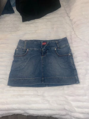 Blå jeanskjol från Kenji Jeans - Säljer en klassisk blå jeanskjol från Kenji Jeans. Kjolen är kort och har fem fickor, bälteshällor och dragkedja framtill. Perfekt för dig som gillar en enkel och tidlös denimlook.