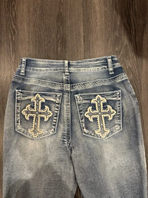 Bootcut jeans med broderade fickor - Snygga ljusblå bootcut jeans med slitningseffekt och broderade kors på bakfickorna. Jeansen har klassisk femficksdesign och utsvängda ben. Perfekta för dig som vill sticka ut med coola detaljer och retrovibbar.