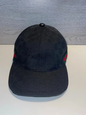 Svart keps från Gucci med logga - Snygg svart keps från Gucci med diskret GG-monogram över hela tyget. Klassisk böjd skärm och ikoniska gröna och röda ränder på sidorna. Tillverkad i slitstark canvas som ger en exklusiv känsla och passar perfekt till streetwear-stilen.