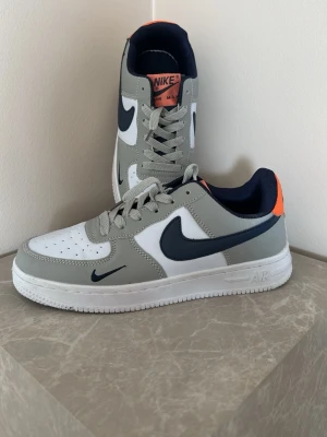 Nike Air Force 1 grå/blå sneakers - Nike Air Force 1 sneakers i grått, vitt och marinblått med orange detalj.  Skorna har grå snörning stl 39