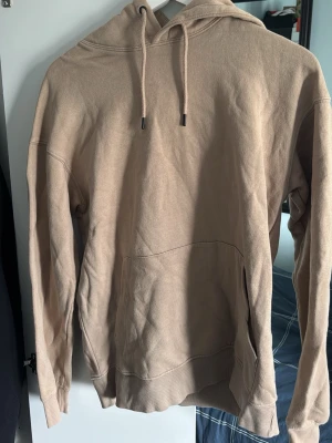 Beige hoodie med ficka framtill - Säljer en beige hoodie med huva och dragsnören. Hoodien har en stor känguruficka framtill och ribbade muddar vid ärmslut och nederkant. Tillverkad i mjukt bomullstyg som är skönt att ha på sig. Perfekt för en chill och avslappnad stil.