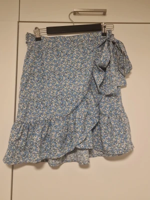 Blommig blå omlottkjol med volang - Söt blå kortkjol med blommigt mönster och volangkant. Kjolen har omlottdesign med en stor knytrosett i sidan och är perfekt för varma dagar. Lätt och luftig känsla, passar till både sneakers och sandaler.