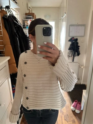 Beige ribbstickad tröja med knappar - Säljer en beige ribbstickad tröja med stora svarta knappar längs sidan och ena axeln. Tröjan har långa, vida ärmar med breda muddar och en avslappnad passform. Perfekt för dig som gillar en unik och trendig look.💗😊