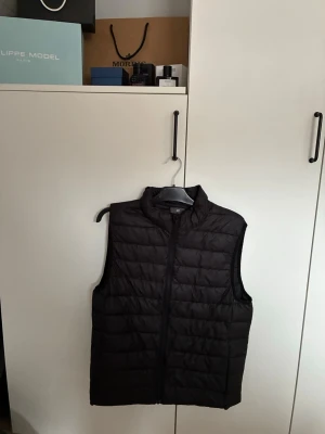 Svart dunväst - Säljer en svart dunväst med dragkedja framtill. Västen har en quiltad design med horisontella sömmar och är ärmlös. Perfekt att slänga över en hoodie eller tröja för extra värme. Enkel och stilren look som funkar till det mesta.