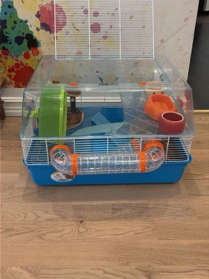 Hamsterbur med tillbehör - Säljer en blå och transparent hamsterbur med vit gallerlucka. Medföljer löphjul, matskålar, vattenflaska, hamsterboll, träkoja, mat, godis och färgglad bottenmatta. Perfekt startkit för hamsterägare, allt du behöver för att komma igång!