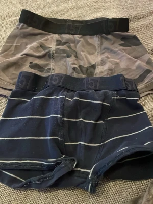 2-pack kalsonger från 157, camo & randig - Två par kalsonger från 157, ena i grått camomönster och den andra i mörkblått med vita ränder. Båda har bred resår med 157-logga. Sköna och stretchiga, perfekta för vardagsbruk. Materialet känns mjukt och följsamt.