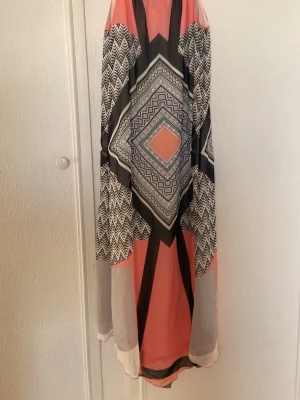 Mönstrad långklänning i korall och svart - Säljer en långklänning med grafiskt mönster i svart, vitt och korall. Klänningen har ärmlös design och ett luftigt, svepande tyg som ger en bohemisk känsla. Perfekt för varma dagar och sommarens alla tillfällen.