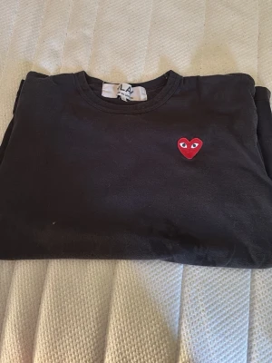 Svart Comme des Garçons t-shirt - Svart t-shirt från Comme des Garçons Play med det ikoniska röda hjärtat med ögon broderat på bröstet. Klassisk rund hals och korta ärmar. Tillverkad i mjuk bomull som känns skön mot huden. Perfekt för dig som gillar stilrena plagg med en twist.