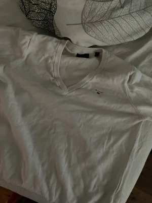 Vit V-ringad tröja från Gant - Snygg vit tröja från Gant med V-ringning och diskret logga broderad på bröstet. Tröjan har långa ärmar och ribbade muddar vid hals, ärmslut och nederkant. Perfekt till jeans eller kjol för en clean och klassisk look.