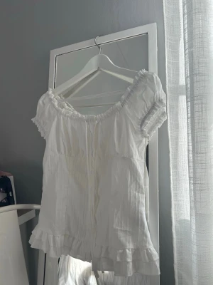 Vit blus med volang och knappar - Söt vit blus med korta puffärmar, volangkant nedtill och knappar framtill. Blusen har en lätt rynkad resår vid halsringningen och ärmarna, vilket ger en romantisk vibe. Tillverkad i ett tunt, luftigt bomullstyg som passar perfekt till sommaren.