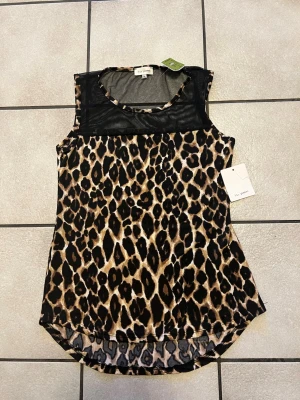 Leopardmönstrad ärmlös topp Blu Planet storlek M - Snygg ärmlös topp från Blu Planet med leopardmönster och transparent meshparti upptill. Rund halsringning och längre baktill för en cool look. Perfekt för dig som vill sticka ut med djurmönster och trendiga detaljer.
