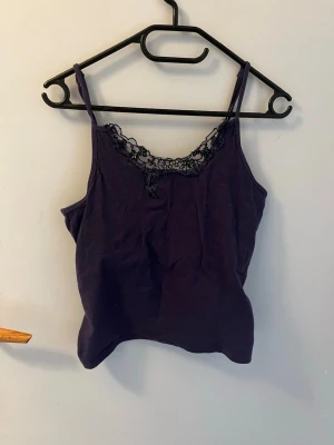 Mörklila linnetopp med spets - Snygg mörklila linnetopp med tunna axelband och dekorativ spetskant vid urringningen. Spetsen har små svarta pärlor som ger en extra detalj.  Från secret possessions. 