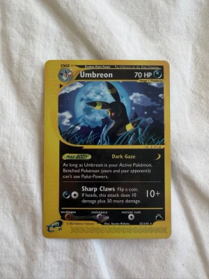 Pokemonkort Umbreon skryridge 32/144 - Säljer ett samlarkort från Pokémon, Umbreon e-Card 32/144. Kortet har en svart och gul design med motiv av Umbreon mot en nattlig bakgrund. Text och symboler i silver och gult. Perfekt till samlingen eller för Pokémon-fans.