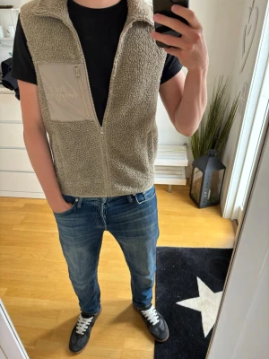 Beige fleeceväst från Peak Performance - Mysig beige fleeceväst från Peak Performance med dragkedja framtill och hög krage. Västen har en bröstficka i nylon med broderad logga och två sidofickor. Perfekt för lager-på-lager och chill stil.