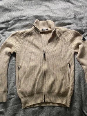 Beige stickad ziptröja med hög krage - Säljer en beige stickad ziptröja med hög krage och två fickor med dragkedja. Tröjan har ribbade muddar och en svart patch på ärmen. Perfekt för lager på lager och chill stil.