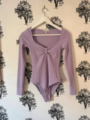 Ljuslila ribbad body med lång ärm - Supersnygg ljuslila body med långa ärmar och ribbad struktur. Djup, rundad ringning framtill med en söt twistdetalj vid bysten. Stretchigt material som sitter tajt och bekvämt. Perfekt att styla med jeans eller kjol för en trendig look.