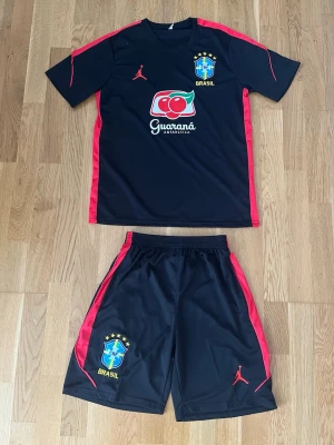 Brasilien träningsset Jordan svart/röd - Snyggt träningsset från Brasilien med Jordan-logga. Setet består av en svart t-shirt och shorts med röda detaljer längs sidorna. Både tröjan och shortsen har Brasilien-emblem och Guaraná Antarctica-tryck. Tillverkat i lätt och snabbtorkande material, perfekt för träning.