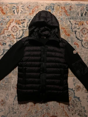 Moncler cardigan  - Snygg svart dunjacka från Moncler med quiltad front och huva. Jackan har släta svarta ärmar och en Moncler-logga på vänster ärm. Perfekt för kalla dagar med sin varma dunfyllning och stilrena design.