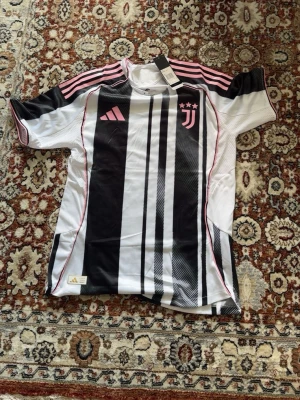 Juventus matchtröja Adidas svartvit - Juventus officiella matchtröja från Adidas i svartvitt med rosa detaljer och tre stjärnor på bröstet. Tröjan har korta ärmar, klassiska Adidas-ränder på axlarna och är tillverkad i lätt, ventilerande material perfekt för träning eller match.