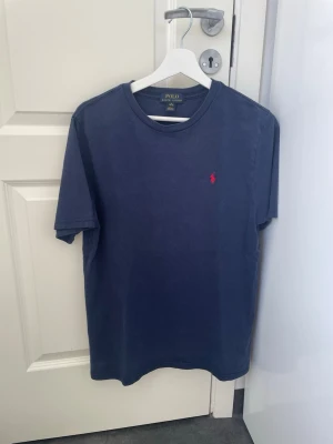 Blå t-shirt från Polo Ralph Lauren - Klassisk marinblå t-shirt från Polo Ralph Lauren med röd broderad logga på bröstet. T-shirten har rund halsringning och korta ärmar. Tillverkad i mjuk bomull för en skön och avslappnad stil.