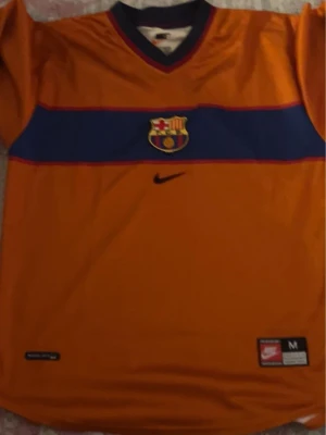 Barcelona matchtröja orange Nike - Snygg orange FC Barcelona matchtröja från Nike med blått och rött band över bröstet och klubbmärke i mitten. Tröjan har korta ärmar, V-ringad krage med blå och röda detaljer och är tillverkad i ett lätt funktionsmaterial.