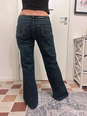 Mörkblå bootcut jeans Lee Cooper - Säljer ett par mörkblå bootcut jeans från Lee Cooper med snygga kontrastsömmar och broderade bakfickor. Jeansen har lågmidjade och klassisk femficksdesign. Perfekta för dig som gillar en avslappnad men trendig look.Midja: 39 Höft: 41 Gren: 19 Lår: 26 Innerben: 75 Benvidd (längst ner): 26
