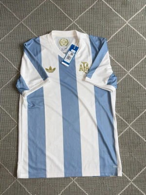 Argentina Adidas retro fotbollströja - Säljer en klassisk Argentina retro fotbollströja från Adidas med blå och vita vertikala ränder, guldfärgade detaljer och AFA-logga på bröstet. Tröjan har v-ringning och är tillverkad i ett lätt, ventilerande material. Perfekt för dig som älskar fotboll och snygg sportstil. Helt ny aldrig använt ny pris 1099kr