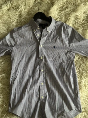 Randig skjorta från Ralph Lauren - Klassisk blåvit randig skjorta från Ralph Lauren med button-down krage och broderad logga på bröstet. Skjortan har långa ärmar och vita knappar framtill. Tillverkad i bomull för en skön och stilren look.
