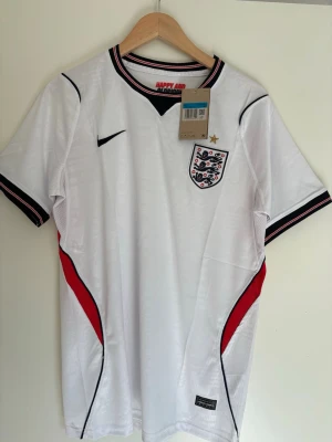 England fotbollströja Nike Dri-FIT - Snygg vit England fotbollströja från Nike med Dri-FIT material. Tröjan har svarta och röda detaljer, broderat landslagsemblem med stjärna, samt texten 'HAPPY AND GLORIOUS' i nacken. Perfekt för match eller träning.
