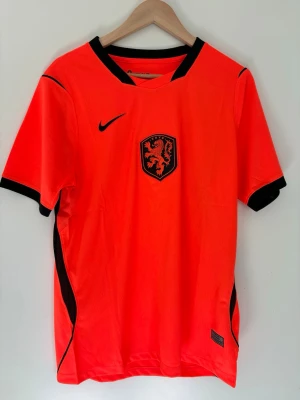 Nederländerna  Nike fotbollströja orange - Snygg orange fotbollströja från Nike med svarta detaljer och Holland-lejonet på bröstet. Tröjan har korta ärmar, svart krage och är tillverkad i lätt, ventilerande Dri-Fit material. Perfekt för träning eller match!