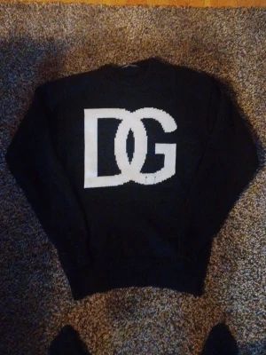 Svart stickad tröja Dolce & Gabbana - Snygg svart stickad tröja från Dolce & Gabbana med stor vit DG-logga framtill. Klassisk rund halsringning och ribbade muddar vid ärmslut och nederkant. Perfekt för dig som gillar exklusiva och trendiga plagg.