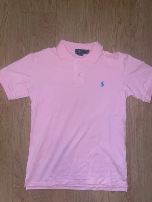 Rosa pikétröja från Polo Ralph Lauren - Säljer en klassisk rosa pikétröja från Polo Ralph Lauren med blå broderad logga på bröstet. Tröjan har krage, korta ärmar och knäppning framtill. Perfekt till en avslappnad men ändå stilren look.