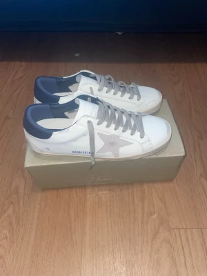 Golden Goose Superstar vita sneakers - Golden Goose Superstar sneakers i vitt läder med grå stjärndetalj på sidan, grå skosnören och mörkblå häl. Klassisk låg modell med perforerade detaljer och märkning på insidan. Snygg och tidlös design som passar till det mesta.