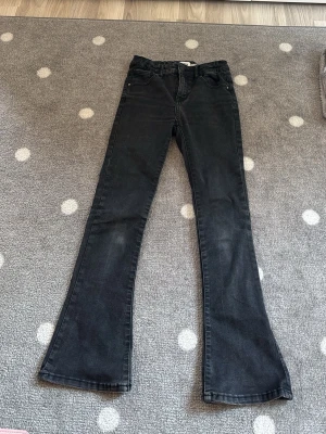 Svarta bootcut jeans från KappAhl - Snygga svarta bootcut jeans från KappAhl med klassisk femficksmodell och hög midja. Jeansen har utsvängda ben och är tillverkade i ett mjukt denimtyg med lite stretch för extra komfort. Perfekta för dig som gillar en trendig och avslappnad stil.