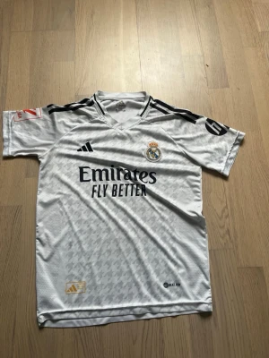 Real Madrid matchtröja Mbappé 9 - Snygg vit Real Madrid matchtröja från Adidas med svarta detaljer och tryck. På framsidan finns klubbmärket och sponsorlogga, på ryggen står det Mbappé och nummer 9. Tillverkad i lätt och ventilerande material, perfekt för fotbollsträning eller match.