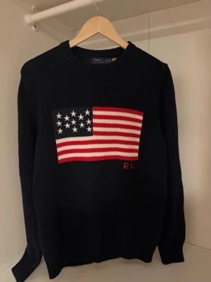 Mörkblå stickad tröja Polo Ralph Lauren - Stickad mörkblå tröja från Polo Ralph Lauren med stor amerikansk flagga i rött, vitt och blått på bröstet samt RL-broderi. Tröjan har rund halsringning och ribbade muddar. Perfekt för dig som gillar klassisk preppy stil.