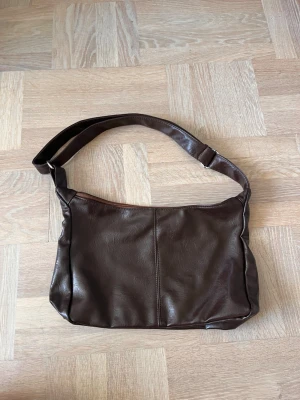Mörkbrun axelväska i fejkläder - Snygg mörkbrun axelväska i fejkläder med enkel, stilren design. Väskan har justerbart axelband och dragkedja upptill. Perfekt storlek för att få plats med det viktigaste och passar till många olika outfits.