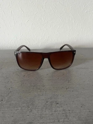 solglasögon - Säljer ett par stilrena solglasögon med bred, mörkbrun båge och stora rektangulära glas. Glasen har en snygg tonad brun färg som ger en cool vibe. Perfekta för soliga dagar och ger en modern look till din outfit.
