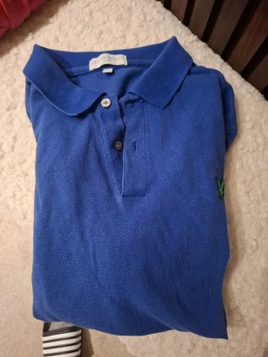 Blå pikétröja från Lyle & Scott - Klassisk blå pikétröja från Lyle & Scott med krage och knappar framtill. Tröjan har en liten broderad logga i grönt på bröstet och är tillverkad i ett bekvämt bomullsmaterial. Perfekt till jeans eller shorts.