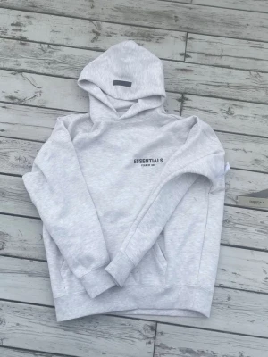 Grå Essentials Fear of God hoodie - Snygg ljusgrå hoodie från Essentials Fear of God med svart logga tryckt på bröstet och ryggen. Hoodien har en klassisk huva, ribbade muddar och är gjord i mjukt bomullsmaterial. Perfekt för en avslappnad och trendig streetwear-look.