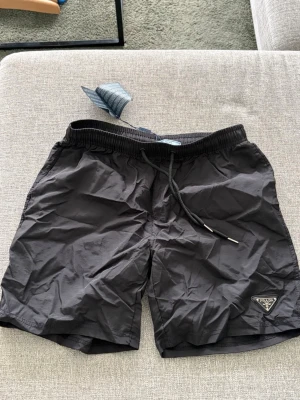 Svarta badbyxor från Prada - Snygga svarta badbyxor från Prada med elastisk midja och dragsko för perfekt passform. Klassisk design med diskret Prada-logga på vänster ben. Tillverkade i lätt och snabbtorkande material, perfekta för stranden eller poolen.
